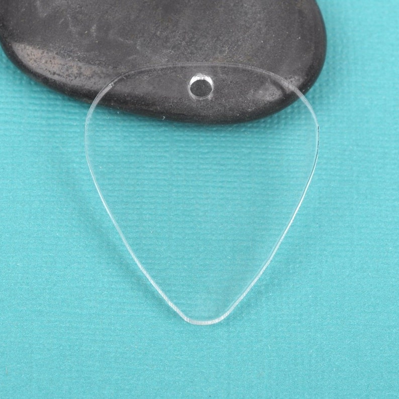 May include: A clear plastic guitar pick with a hole at the top for a string or chain. The pick is shaped like a traditional guitar pick and is on a blue background.