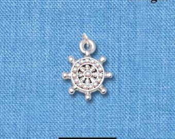 May include: A silver-colored charm in the shape of a ship's wheel, with a central design and surrounding spokes. The charm is approximately 0.5 inches in diameter. The word "Delight" is printed in the upper right corner. The charm is on a blue textured background.