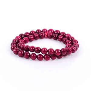 May include: A double strand of round, polished beads in a deep burgundy color. The beads are strung together, forming a circular shape. The jewelry is set against a white background.