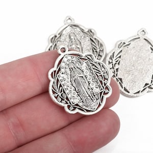 May include: Silver-toned metal pendant with a religious figure in the center, surrounded by a floral filigree design. The pendant has a small loop at the top for attaching to a chain or cord. The image shows multiple pendants.