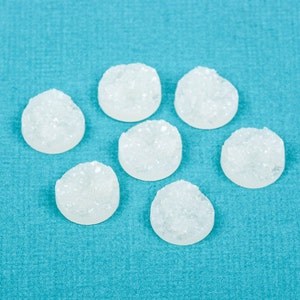 May include: Seven round, white, crystalline objects are arranged on a textured, turquoise blue surface. The objects have a rough, textured appearance, and are clustered together. The background is a solid, light blue color.