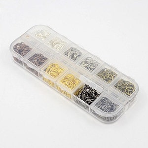 Op de afbeelding: Een doorzichtige plastic organizer gevuld met diverse kleuren verbindingsringen. De verbindingsringen zijn in tinten zilver, goud, brons en zwart. De organizer heeft 12 compartimenten.