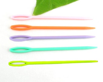 May include: A collection of five colorful plastic yarn needles in pink, orange, purple, teal, and green. Each needle has a rounded eye and a tapered point. A green leaf is in the background.