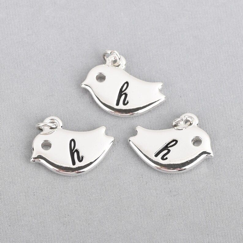 May include: Three silver bird charms, each with a small loop for hanging. The charms have a smooth, reflective surface and feature the lowercase letter "h" in black. The birds are stylized with a simple, rounded shape.