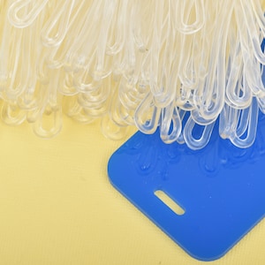 May include: A collection of clear plastic loop fasteners and a blue rectangular tag with a slot. The fasteners are clustered together, and the tag is positioned at an angle on a yellow surface.
