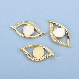 May include: Three gold-colored eye-shaped pendants. Each pendant features a white opal-like center and is outlined with small, clear crystals. The pendants have a loop at each end for attaching to jewelry.