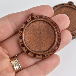 Op de afbeelding: Ronde houten hangerbasis met een centraal verzonken gebied en decoratieve rand. De rand heeft afwisselend kleine cirkels en bloemvormige uitsparingen. De hanger heeft een kleine lus aan de bovenkant om op te hangen. Het hout heeft een warme, roodbruine tint.