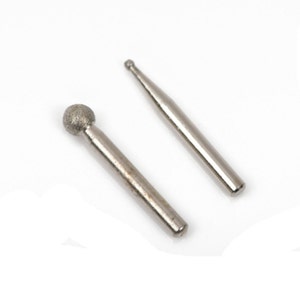 May include: Two silver-colored rotary burrs on a white background. One burr has a ball-shaped head with a textured surface, and the other has a pointed tip. These tools are used for grinding, shaping, and smoothing materials.
