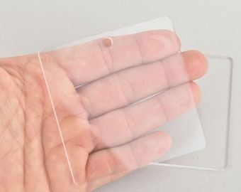 May include: Clear, rectangular acrylic blanks with rounded corners and a small hole for hanging. One blank is held in a hand, with another lying beneath it. The transparent material allows light to pass through, creating a clean, modern look.