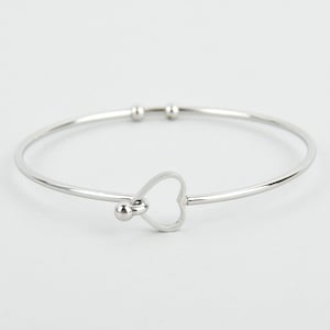 Puede incluir: Una pulsera de brazalete plateada con un dije en forma de corazón. La pulsera tiene un diseño simple y elegante con una banda delgada y un pequeño corazón abierto en el centro. La pulsera está hecha de un metal brillante y reflectante.