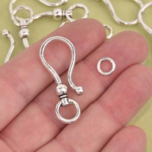May include: Silver-colored metal jewelry findings, including a hook clasp with a round ring and a small jump ring. The hook clasp has a curved design with a ball at the end of the hook.