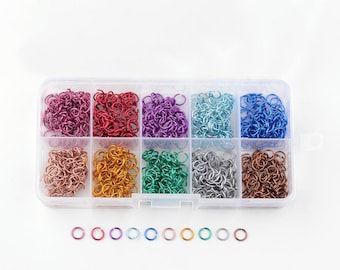 May include: A clear plastic storage box filled with small metal jump rings in various colors, including pink, purple, blue, green, silver, gold, and copper. The box has eight compartments, each containing a different color of jump rings. There are also individual jump rings in each color lined up below the box.