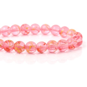 May include: A bracelet featuring translucent pink beads with gold-colored vein-like patterns. The round beads are strung together, creating a delicate and elegant accessory. The bracelet is set against a white background.