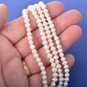 May include: A close-up of three strands of small, creamy white pearls. The pearls are strung together, creating a delicate necklace or bracelet. The background is a solid blue color.