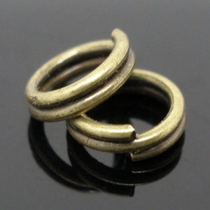 May include: Two antique brass-colored split rings, one partially open, are stacked on a reflective surface. The rings have a double-loop design, commonly used in jewelry making and crafts. The rings are made of metal.