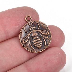 May include: A copper-colored metal charm with a detailed bee design. The charm is round and has a loop at the top for attaching to a necklace or bracelet.