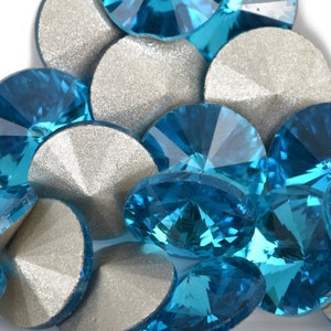 May include: A close-up of a pile of turquoise blue rhinestones. The rhinestones are round and have a faceted surface. They are scattered on a silver background.