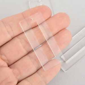 May include: Clear, rectangular acrylic earring blanks with a cutout rectangle and a small hole at the top for jewelry making. Several blanks are visible, held in a hand.