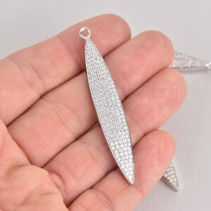 May include: Silver-toned, elongated pendant with a leaf-like shape, densely covered in small, sparkling crystals. The pendant has a small loop at the top for attaching to a chain or other jewelry component. Additional pendants are visible in the background.