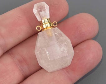May include: A clear, faceted crystal perfume bottle with a gold-colored metal collar and stopper. The bottle has a geometric shape and is held in a hand against a gray background. The stopper is also crystal and faceted.