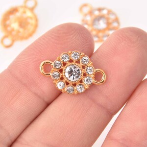 Puede incluir: Conector de joyería dorado con diseño floral. El conector presenta un gran cristal transparente en el centro, rodeado de cristales más pequeños. Dos bucles a cada lado para la fijación. Otros conectores son visibles en el fondo.