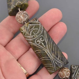 May include: Close-up of a jade necklace with carved rectangular and spherical beads. The jade is a dark green color with intricate white etched designs. The necklace is held in a hand against a gray background.