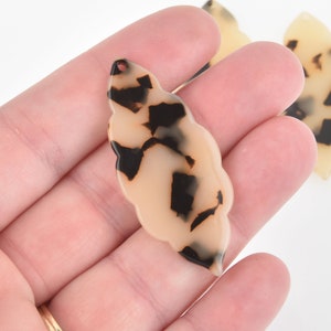 May include: A collection of leaf-shaped pendants in a tortoiseshell pattern. The pendants are a light beige color with dark brown and black spots. Each pendant has a small hole at the top for stringing.
