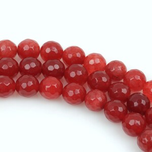 May include: Strands of faceted red gemstone beads. The beads have a polished, reflective surface and are arranged in curved rows against a white background. The beads vary in shade from a deep burgundy to a bright red.