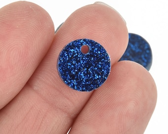 May include: Close-up of a small, round, blue glitter pendant with a hole at the top. The pendant is held between fingers against a white background. The glitter gives the pendant a sparkling appearance.