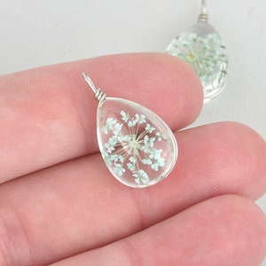 May include: Clear teardrop-shaped pendant with a silver wire loop, encasing delicate, light blue dried flowers. The pendant is held in a hand, with another similar pendant visible in the background. The jewelry has a natural, botanical theme.