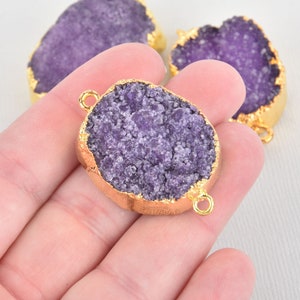 May include: Close-up of several purple druzy gemstone pendants with gold-colored edges. The pendants have a rough, textured surface and are oval-shaped with small loops for attaching to jewelry. The background is a light, neutral color.