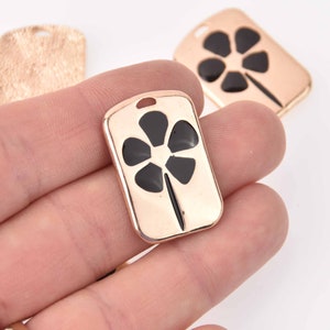May include: Gold-tone metal charm with a black enamel flower design. The charm is rectangular in shape and has a hole at the top for attaching to a necklace or bracelet.