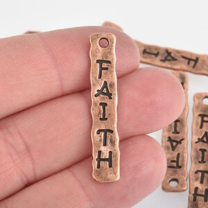 May include: Copper-colored metal charm with the word "FAITH" vertically stamped in black. The charm has a textured, hammered appearance and a small hole at the top for stringing. Several similar charms are visible in the background.