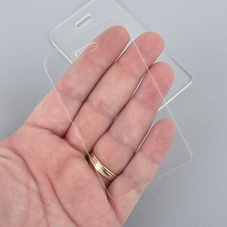 Puede incluir: Dos porta-identificaciones de pl&aacute;stico transparente. Los porta-identificaciones son rectangulares con esquinas redondeadas y un agujero en la parte superior para un cord&oacute;n. Los porta-identificaciones est&aacute;n apilados uno encima del otro.