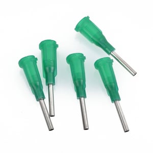 May include: Five green and silver medical needles are scattered on a white surface. The needles have a green plastic base and a silver metal tip. The needles are angled in different directions.
