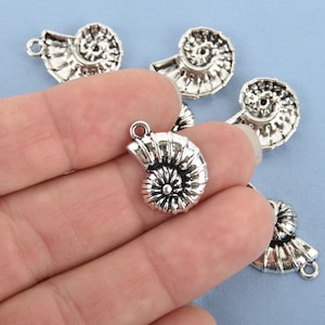 May include: Silver-toned metal charms in the shape of nautilus shells. Each charm has a loop at the top for attaching to jewelry or other crafts. The charms are detailed with spiral ridges and a textured surface.