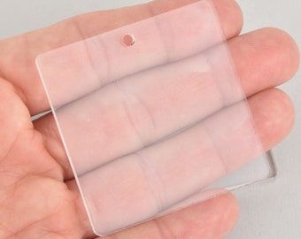May include: A clear, square-shaped acrylic tag with a small hole at the top. The tag is held in a hand, showcasing its transparency and smooth surface. Ideal for crafting or labeling.
