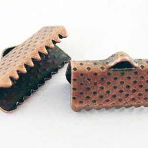 May include: Two copper-colored metal ribbon end crimps. One is open, displaying the interior, and the other is closed. Both have a textured surface with a repeating dot pattern and a serrated edge. They are designed to secure ribbon ends.