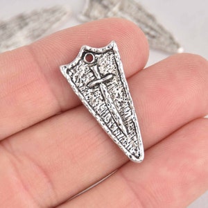 May include: A silver-colored shield charm with a sword design. The shield has a textured surface and a small hole at the top. The sword is positioned vertically through the center of the shield. The charm is held in a hand.
