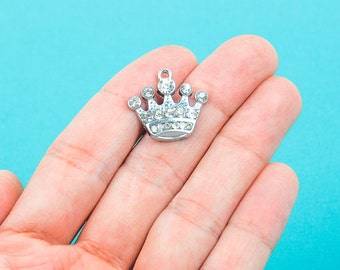 May include: A silver-colored crown charm with clear rhinestones. The charm has a loop at the top for attaching to jewelry. The crown is set against a light blue background.