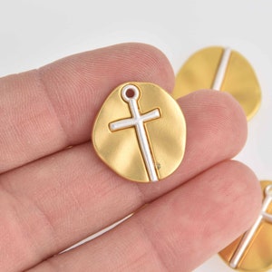Puede incluir: Encanto de metal dorado con un diseño de cruz blanca. El encanto tiene una forma redonda y un agujero en la parte superior para sujetarlo a un collar o pulsera.
