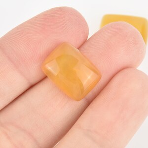 May include: A rectangular, amber-colored gemstone with a polished, glossy surface. The gem is held in a hand, with another similar gem visible in the background. The stone has a warm, translucent appearance.
