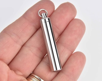 May include: A polished silver cylindrical pendant with a small loop at the top. The pendant is held in a person's hand, showcasing its smooth, reflective surface. The design is simple and elegant, suitable for various styles.