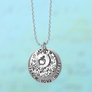 May include: Silver-toned pendant necklace with three circular charms. The largest charm has the words "TEACH LOVE INSPIRE" and a pattern of shapes. The middle charm has an apple graphic. The necklace hangs on a ball chain.