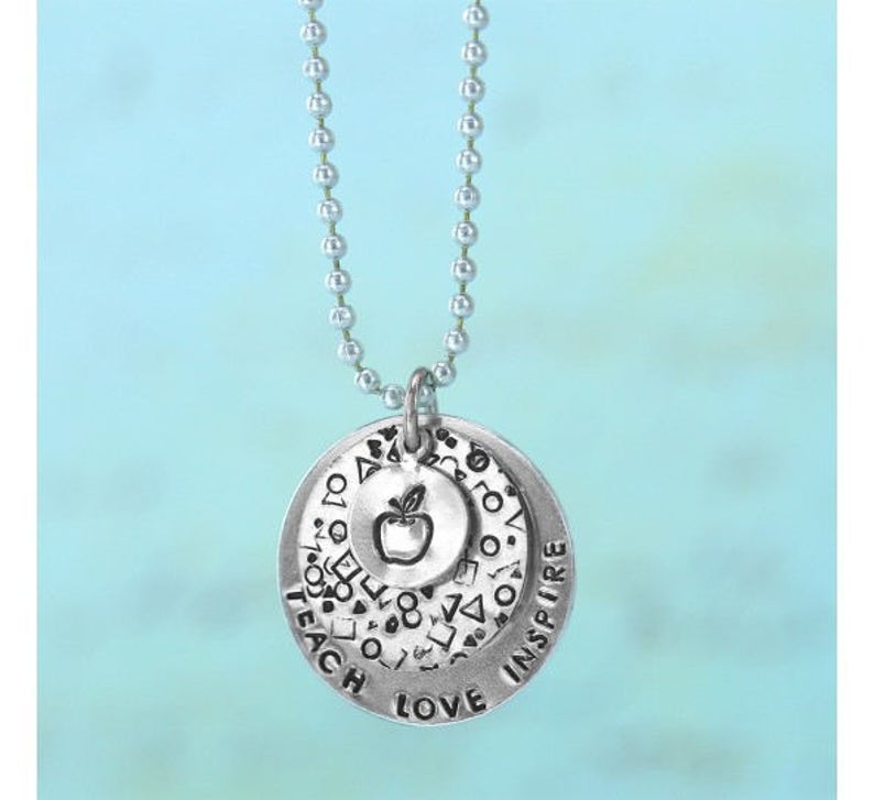 May include: Silver-toned pendant necklace with three circular charms. The largest charm has the words "TEACH LOVE INSPIRE" and a pattern of shapes. The middle charm has an apple graphic. The necklace hangs on a ball chain.