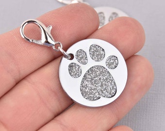 May include: A silver-colored, round pet tag with a paw print design filled with glitter. The tag has a lobster clasp for easy attachment to a collar. The tag is approximately 1 inch in diameter.