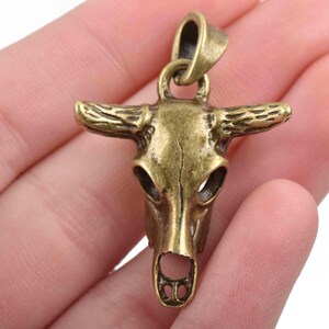 May include: A gold-toned metal pendant in the shape of a bull skull. The pendant has a loop at the top for attaching to a chain or cord.