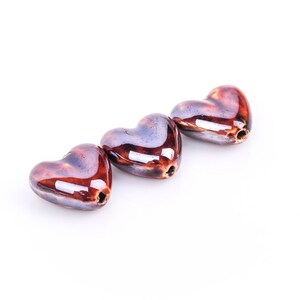 May include: Three heart-shaped beads with a glossy, glazed finish. The beads are a mix of brown and purple hues, with a hole drilled through each for stringing. The beads are arranged in a row against a white background.