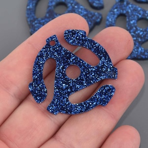 May include: A close-up of several blue glittery vinyl record adapter charms. The charms are in the shape of a 45 RPM record adapter with a circular hole in the center and a small hole at the top. The charms are held in a person's hand.