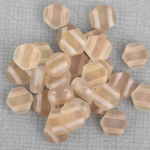 May include: A collection of peach-colored, hexagonal beads with a frosted finish. Each bead features a diagonal stripe design in a lighter shade. The beads are scattered on a gray surface, showcasing their unique shape and color variations.